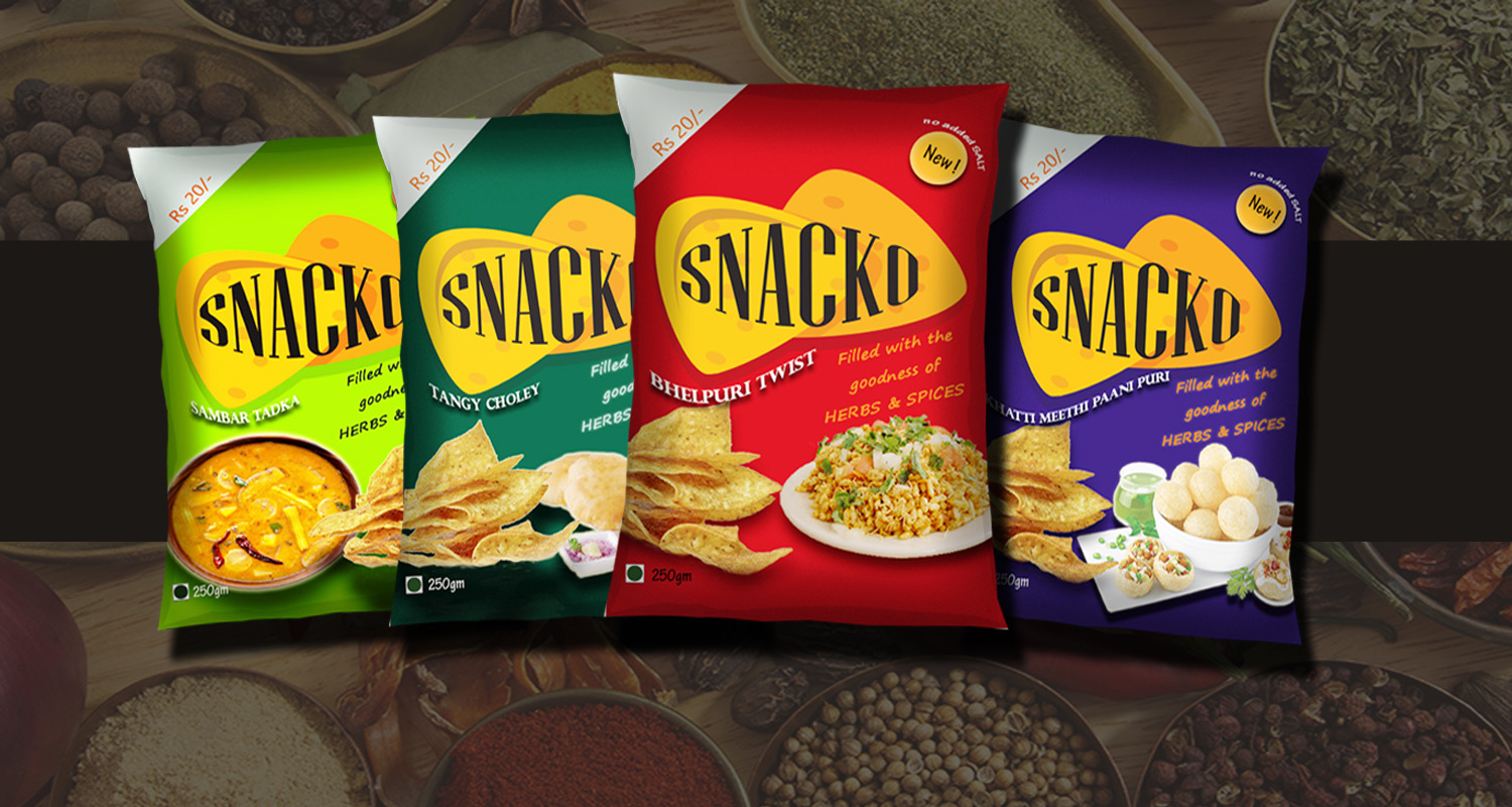 Four Snacko snack packs in green, teal, red and purple flavour variants, lined up on a spice-covered background